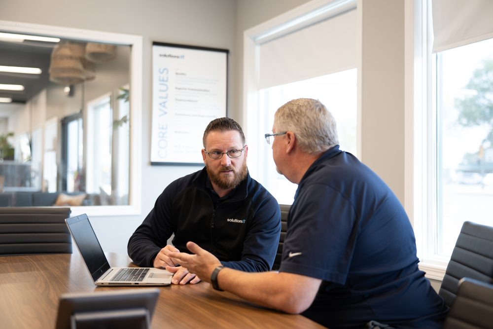Randy at a meeting table in the SolutionsIT office, listening while another man explains something.